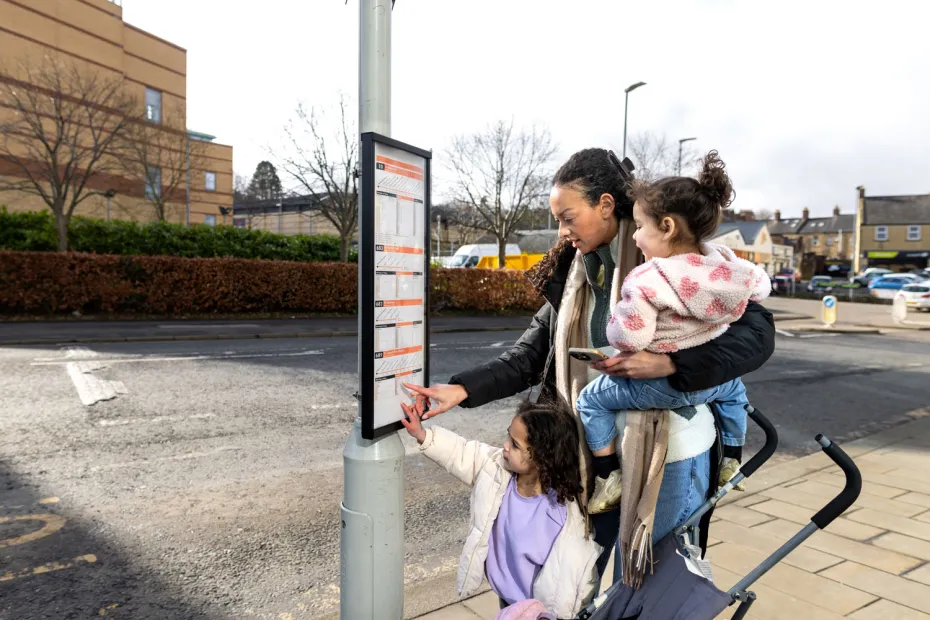 A woman and her two children at a bus stop, looking at the bus timetable