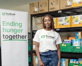 AJ Odudu, Trussell Ambassador, wearing a T-shirt with the Trussell logo standing next to a sign saying ending hunger together