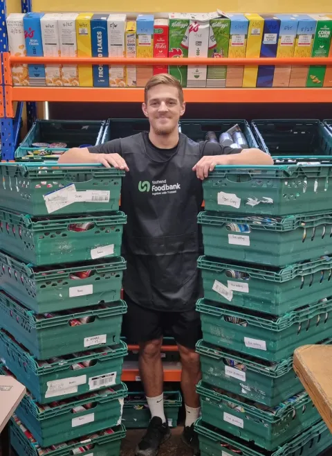 Jack Leech, personal trainer, standing in between two large piles of supermarket crates in a foodbank warehouse.