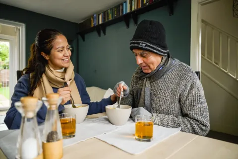 A woman and an elderly man sit at a table with a bowl of food and a glass of apple juice in front of them.
